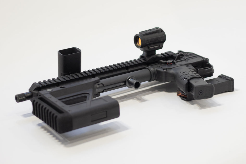 A black tactical rifle accessory setup, including a mounted red dot sight, vertical foregrip in the folded position, and magazine, photographed on a clean white background.