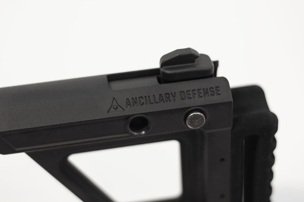 Close-up of a black tactical firearm accessory with the brand name 'ANCILLARY DEFENSE' engraved on the side, set against a plain white background.