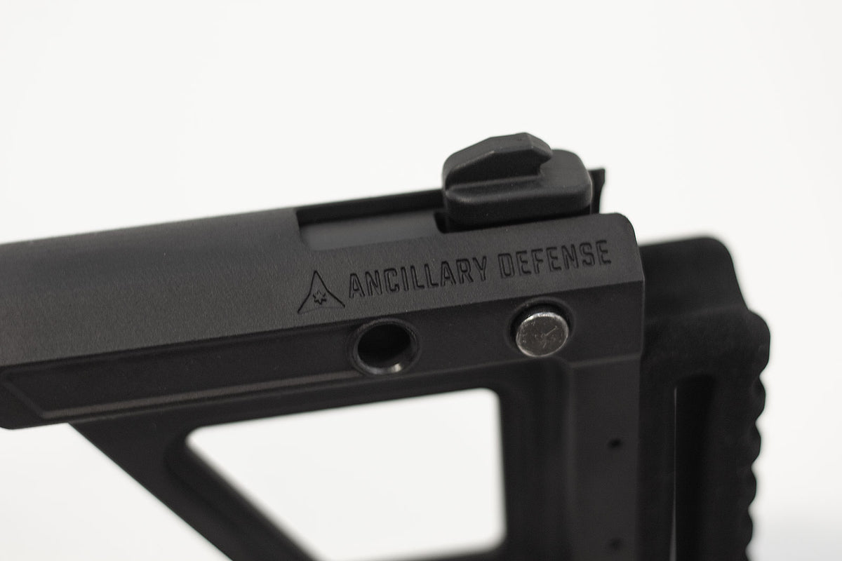 Close-up of a black tactical firearm accessory with the brand name 'ANCILLARY DEFENSE' engraved on the side, set against a plain white background.